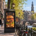 Outdoor burger advertisement beside an Amsterdam canal, with pedestrians, bicycles, and historic buildings in the background.