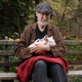 James Cromwell holding a rescued piglet while sitting on a park bench.