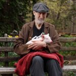 James Cromwell holding a rescued piglet while sitting on a park bench.